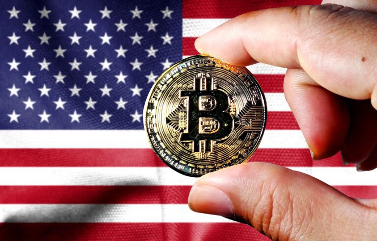 Hand holding a physical Bitcoin with the US flag in the background.