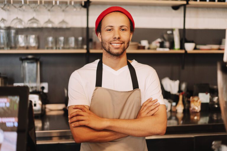 Smiling barista with a red beanie and apron standing in a cafe with crossed arms.