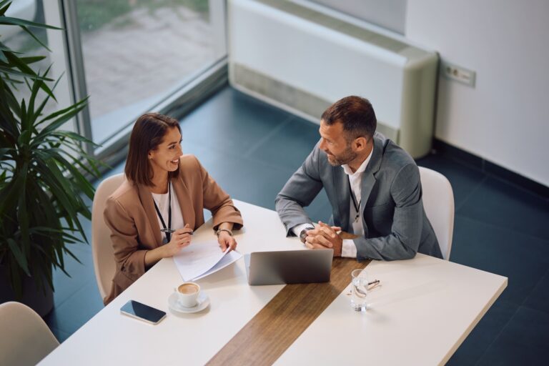 Two professionals having a discussion over a laptop and documents at a white table.