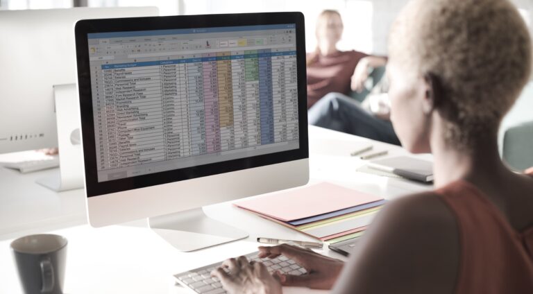 Woman typing on a keyboard while looking at a spreadsheet on a desktop monitor.