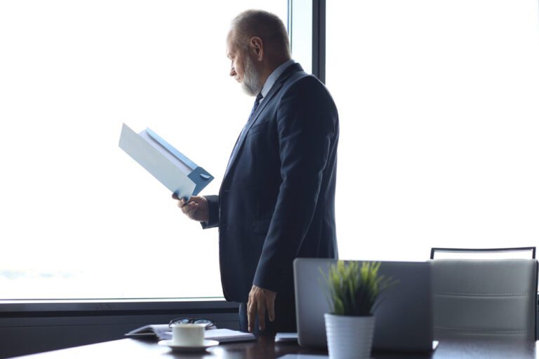 A man in a suit reads a document by a bright window in an office.