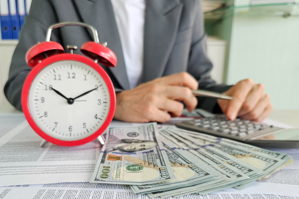 A red alarm clock on a desk next to stacks of US dollar bills and a person using a calculator